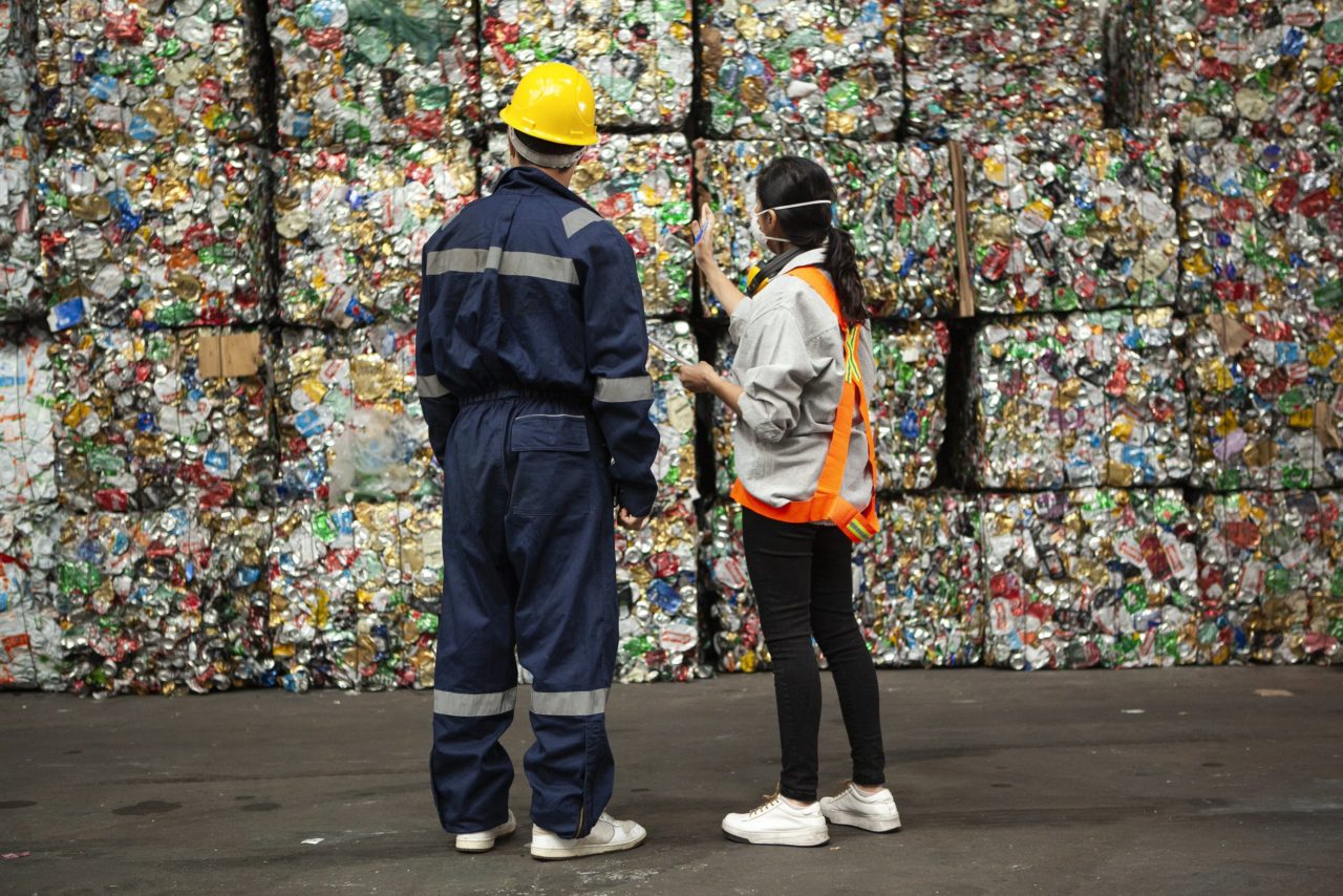Male and female waste management workers standing in front of stacks of compacted recyclables.