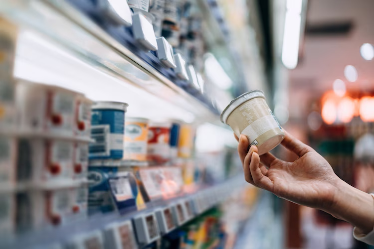 Grocery store shelves full of yogurt options and a single hand selecting one item.