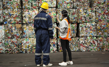 Rear view of male and female waste management workers wearing protective suits and hardhats standing indoors in front of stacks of compacted and bundled recyclables.